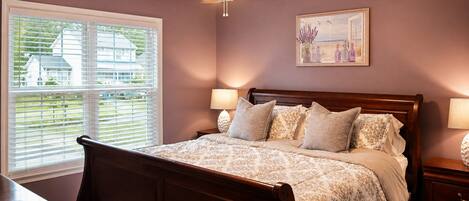 A bedroom with a bed, bedside lamps, a ceiling fan, a window with blinds, and a painting on the wall.