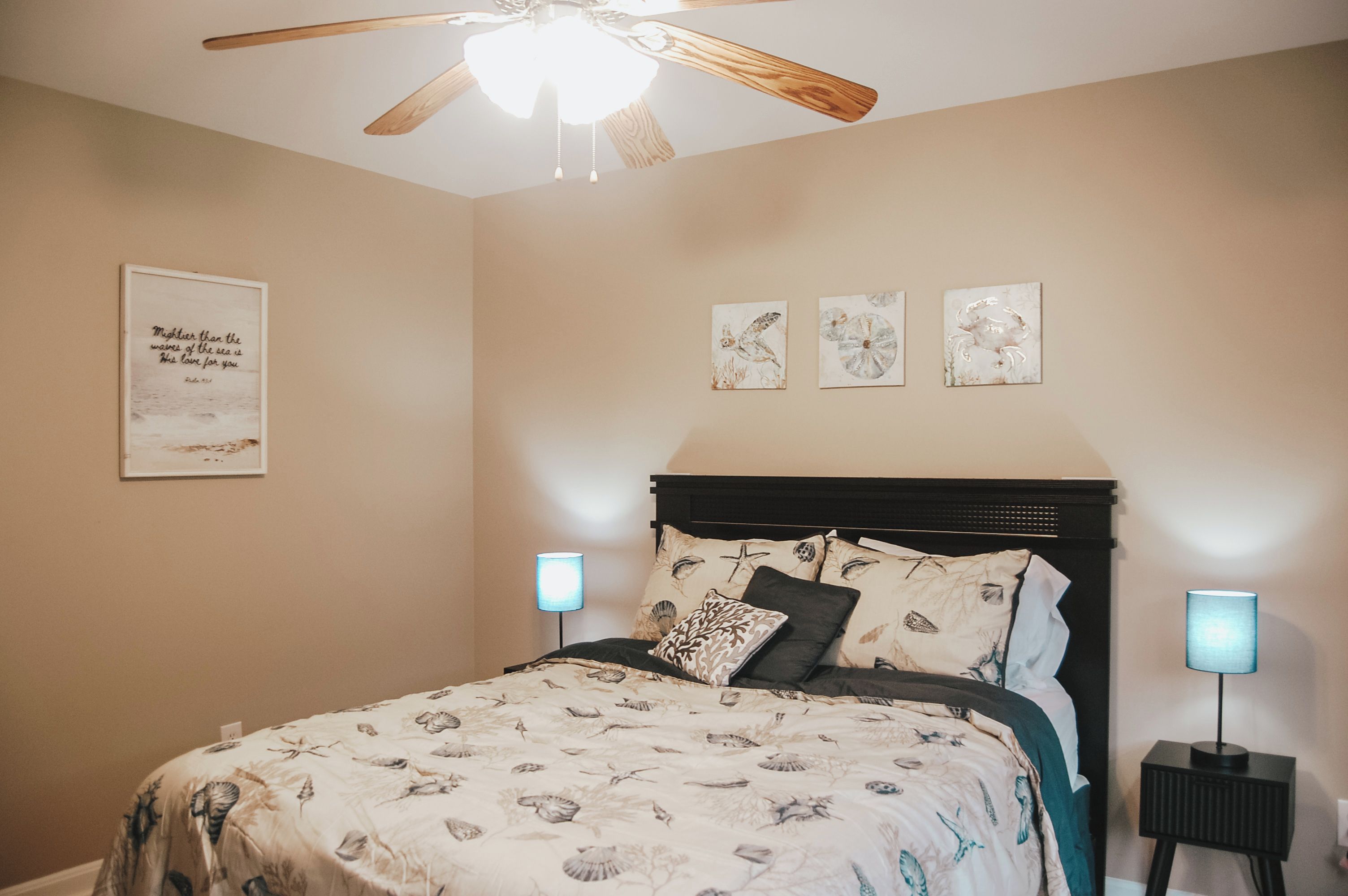 A bedroom with a bed, a ceiling fan, wall-mounted lamps, and three framed artworks.