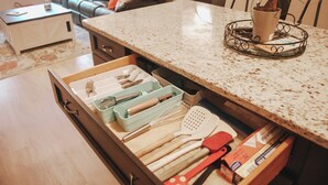 A kitchen with a drawer containing kitchen utensils, a countertop with a plant, and a dining area with a couch and chairs. - Cozy Cottage | Close to Potomac River + Boardwalk (Colonial Beach)