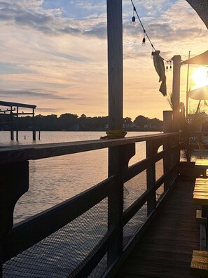 Eine Holzterrasse mit Blick auf ein Gewässer, im Hintergrund ein Sonnenuntergang.