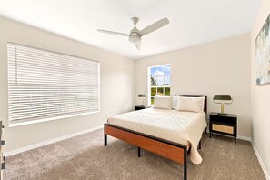 Ein Schlafzimmer mit einem Bett, einem Deckenventilator, einem Nachttisch, einer Lampe und einem Fenster mit Jalousien.