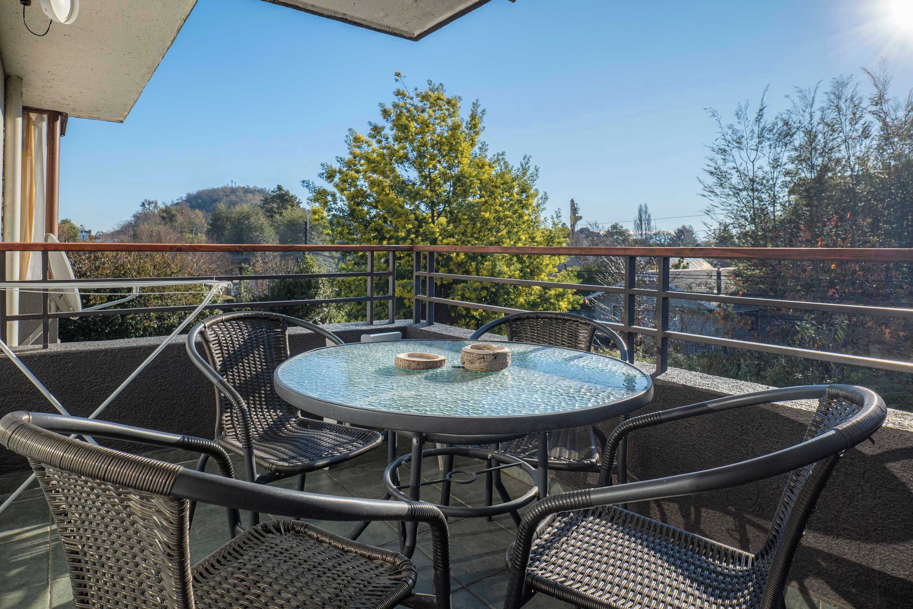 Een balkon met een ronde glazen tafel en rieten stoelen, met uitzicht op een landschap met bomen en heuvels.