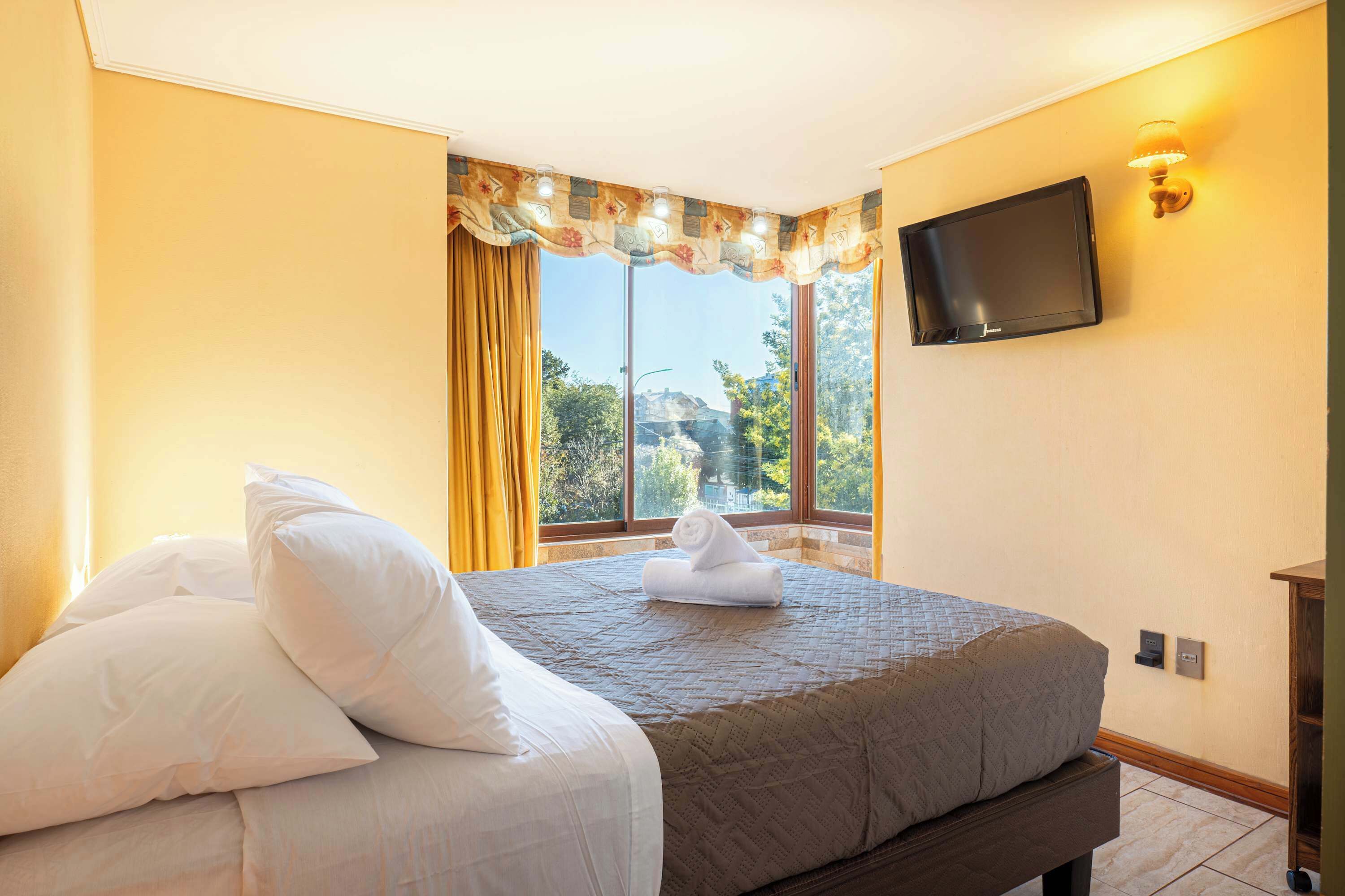 A hotel room with a bed, a flat-screen TV mounted on the wall, and a window with curtains.