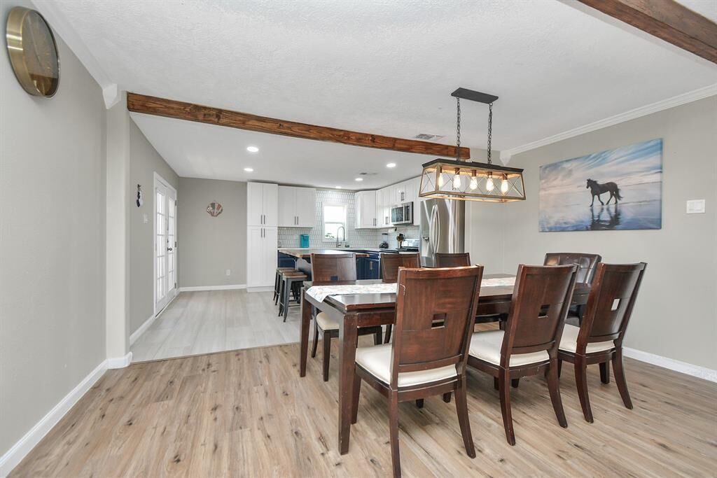 A dining area with a wooden table and chairs, a hanging light fixture, and a kitchen in the background.