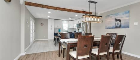 A dining area with a wooden table and chairs, a hanging light fixture, and a kitchen in the background.