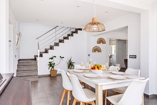 A modern dining area with a white table set for four, surrounded by white chairs, a staircase, and a hanging light fixture.