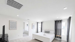 A bedroom with a white bed, a bedside table, a TV stand, and a bathroom visible through an open door.