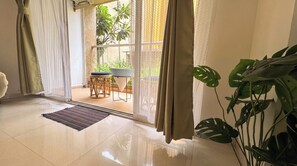 A room with a sliding glass door leading to an outdoor area with a table and chairs, a potted plant, and a large leafy plant in the foreground. - Vibe o1: Boutique 2BHK w/pool | Just 2 mins drive to Anjuna Beach (Anjuna)