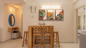 A dining area with a wooden table and chairs, a wall-mounted light fixture, and framed artwork. - Vibe o1: Boutique 2BHK w/pool | Just 2 mins drive to Anjuna Beach (Anjuna)