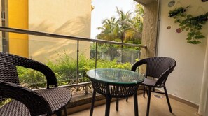 En balkon med et glasbord og to fletstole, med udsigt over en have med forskellige planter og en gul bygning i baggrunden.