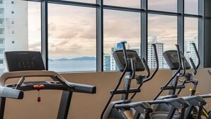 Uma academia com esteiras e uma máquina elíptica, com grandes janelas e vista para a cidade.