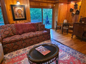 A cozy room with a patterned sofa, a round coffee table with a magazine, and a large window with a view of trees. - Judges Hilltop - Highlands, NC (Highlands)
