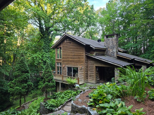 A wooden cabin with a stone chimney, surrounded by dense forest.