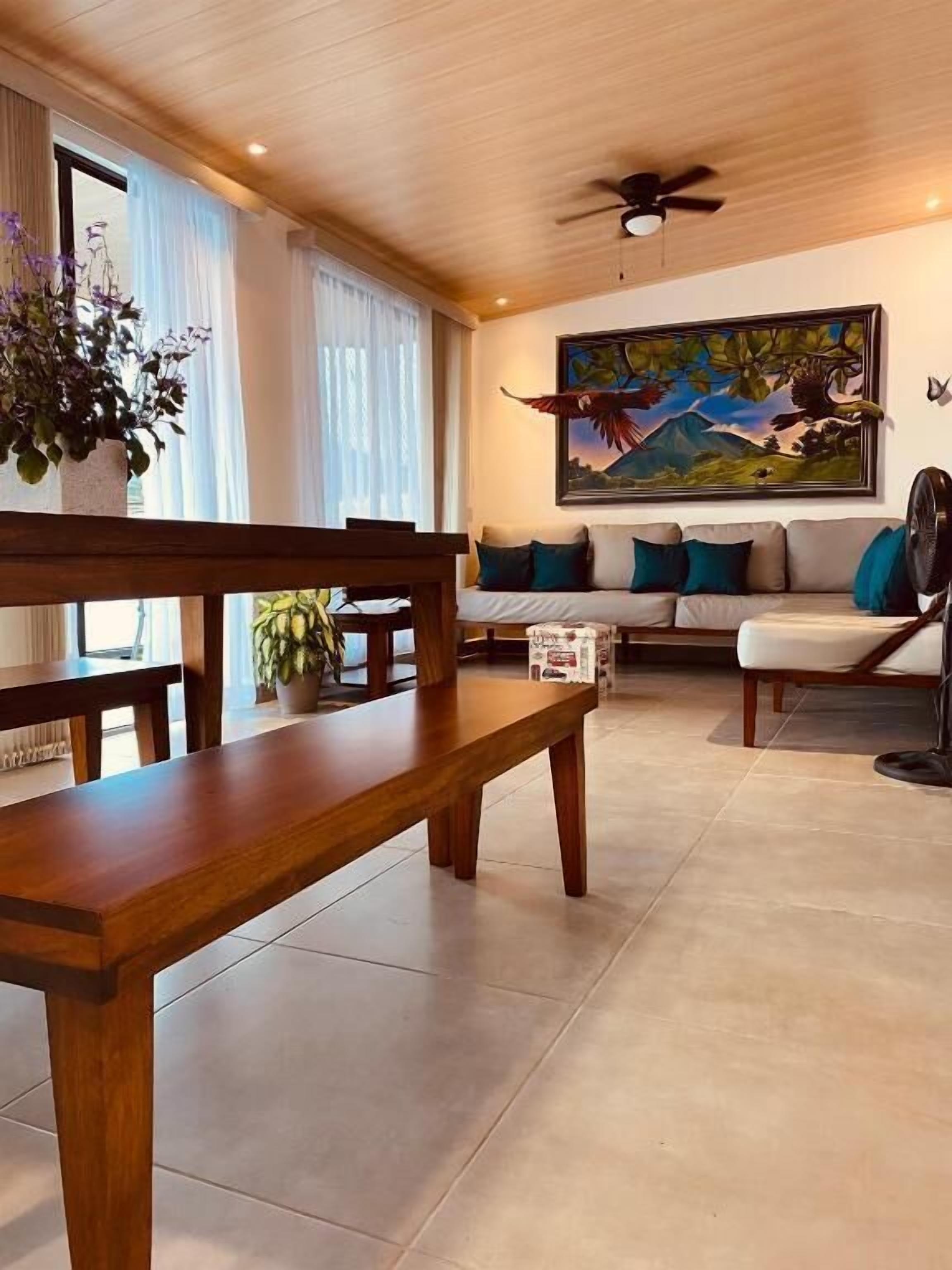 A living room with a wooden bench, a sofa with cushions, a painting on the wall, and a ceiling fan.