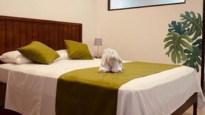 A hotel room with a bed, pillows, a blanket, a small white dog, a potted plant, and a wall-mounted air conditioner.