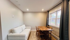 Um quarto com sofá, mesa de jantar de madeira com cadeiras, janela com cortinas e luminária no teto.