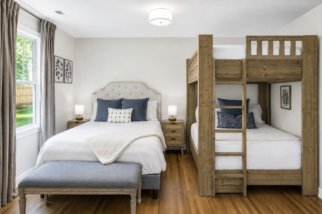 Un dormitorio con una cama grande, una cama más pequeña con escalera de madera, un banco y una mesita de noche con lámpara.