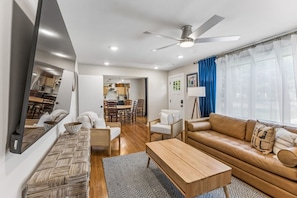 A modern living room with a sofa, coffee table, and chairs, a dining area with a table and chairs, and a kitchen in the background.
