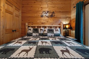 A wooden cabin bedroom with a quilted bedspread featuring animal prints, a decorative headboard, and a nightstand with a lamp. - Orso’s Den, Mountain Views, Theater, Arcade, Hot Tub & Free L2 EV Charging! (Pigeon Forge)