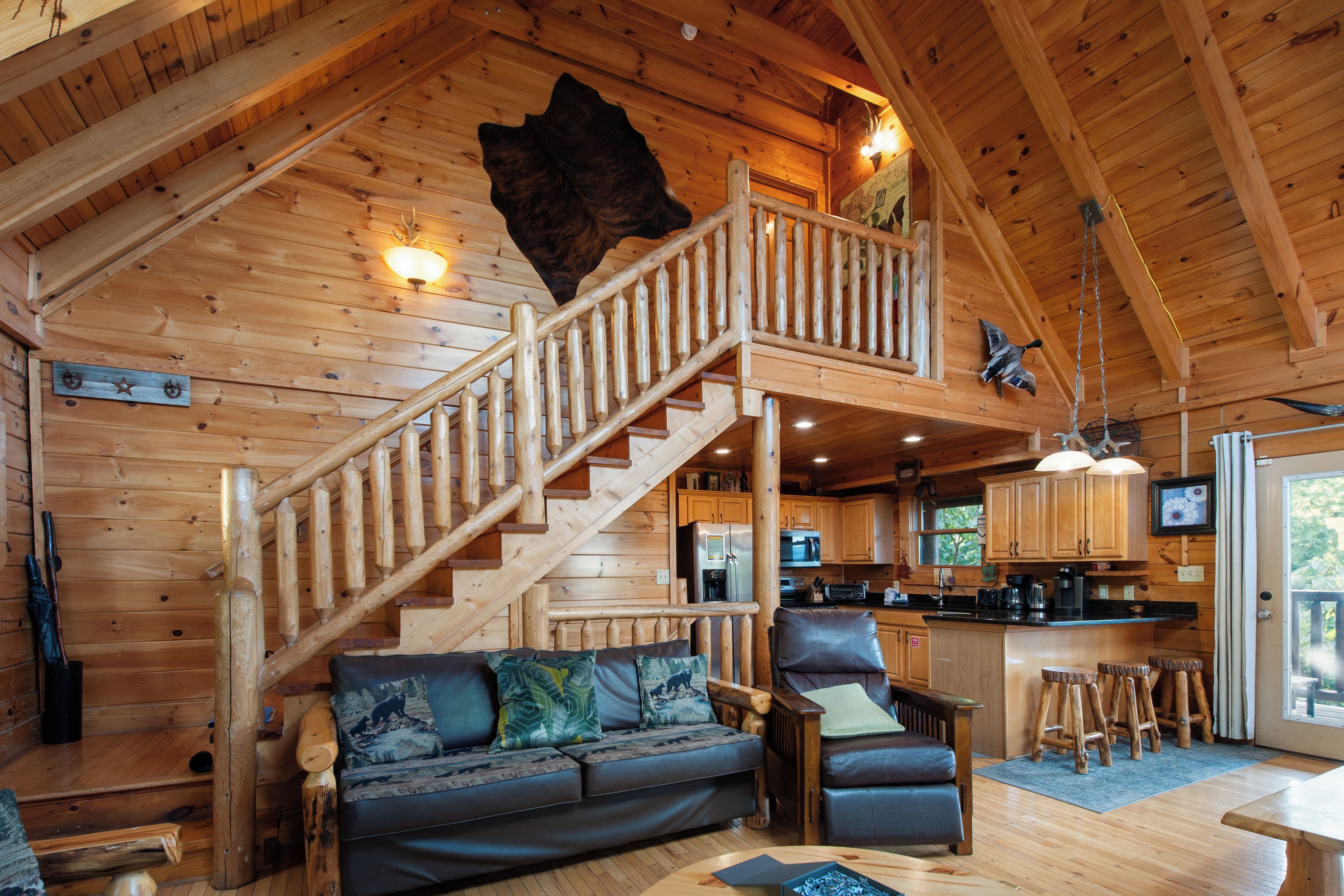 Interior de una cabaña con escalera de madera, sofá, mesa de comedor y zona de cocina.