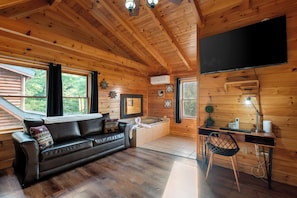 A cabin interior with a wooden ceiling, a black leather sofa, a desk with a computer, and a large TV mounted on the wall. - Orso’s Den, Mountain Views, Theater, Arcade, Hot Tub & Free L2 EV Charging! (Pigeon Forge)