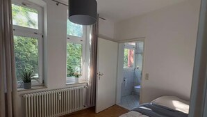 Ett sovrum med en säng, ett element, ett fönster med gardiner och en dörr som leder till ett badrum.