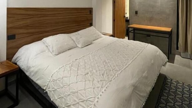 A modern bedroom with a wooden headboard, a large bed with a white bedspread, a bedside table, and a patterned rug. - Casa Valentina (Aguascalientes)