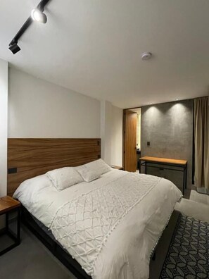 A modern bedroom with a wooden headboard, a large bed with a white bedspread, a bedside table, and a patterned rug. - Casa Valentina (Aguascalientes)