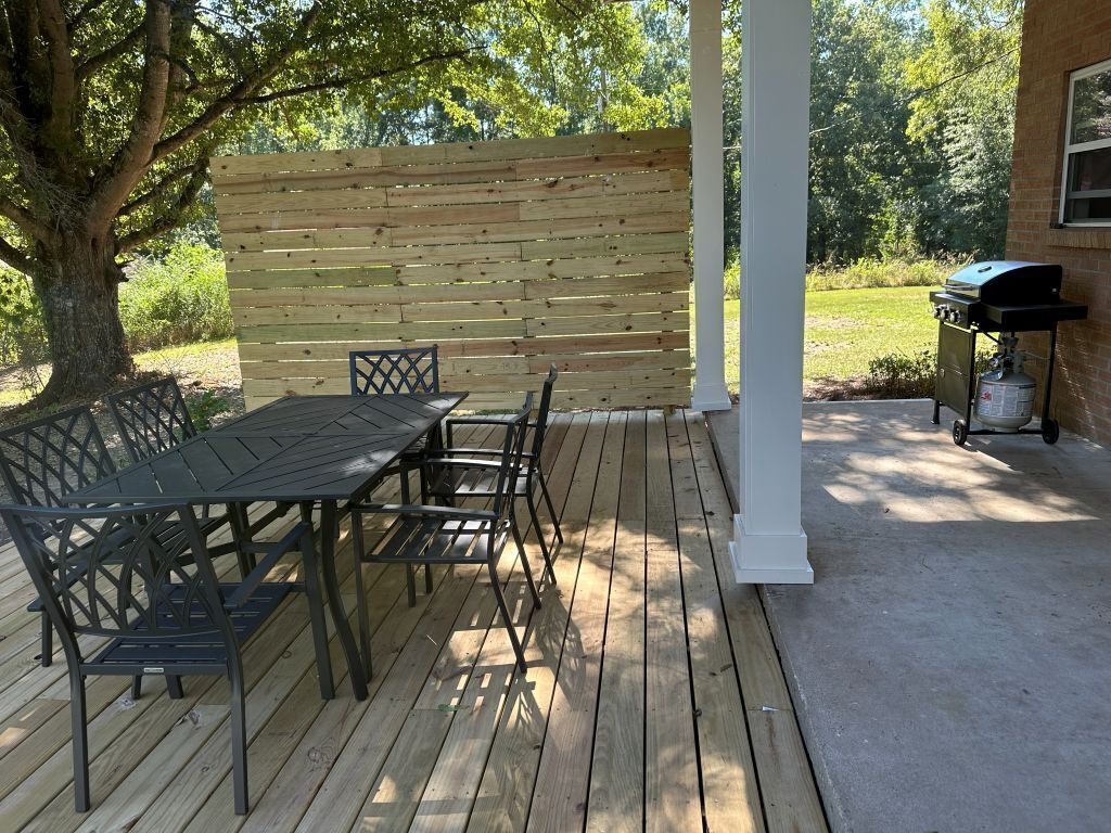 An outdoor patio with a wooden deck, a metal table and chairs, a white column, and a barbecue grill.