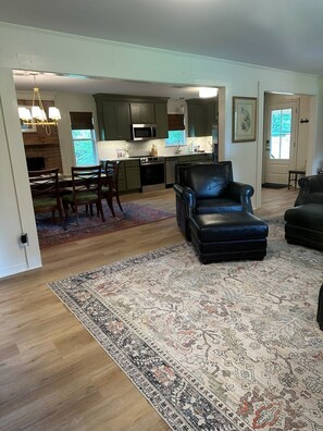 A spacious living room with a large black leather sofa, a dining area with wooden chairs, and a kitchen in the background.