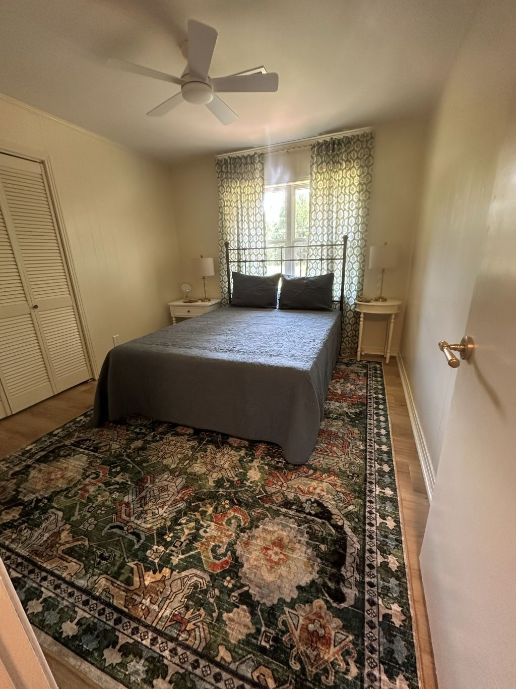 Une chambre à coucher comprenant un lit, un ventilateur de plafond, une fenêtre avec des rideaux à motifs, une table de chevet, une lampe et un tapis décoratif.