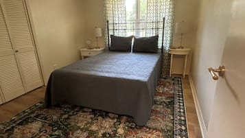 Une chambre à coucher comprenant un lit, un ventilateur de plafond, une fenêtre avec des rideaux à motifs, une table de chevet, une lampe et un tapis décoratif.