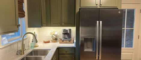 A kitchen with green cabinets, stainless steel appliances, and a window with blinds.