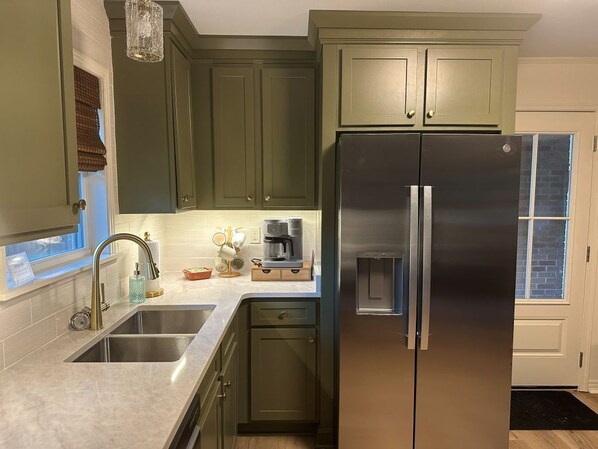 A kitchen with green cabinets, stainless steel appliances, and a window with blinds.