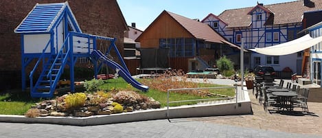 A playground with a blue slide and climbing structure, surrounded by a garden with various plants and a paved area with tables and chairs.