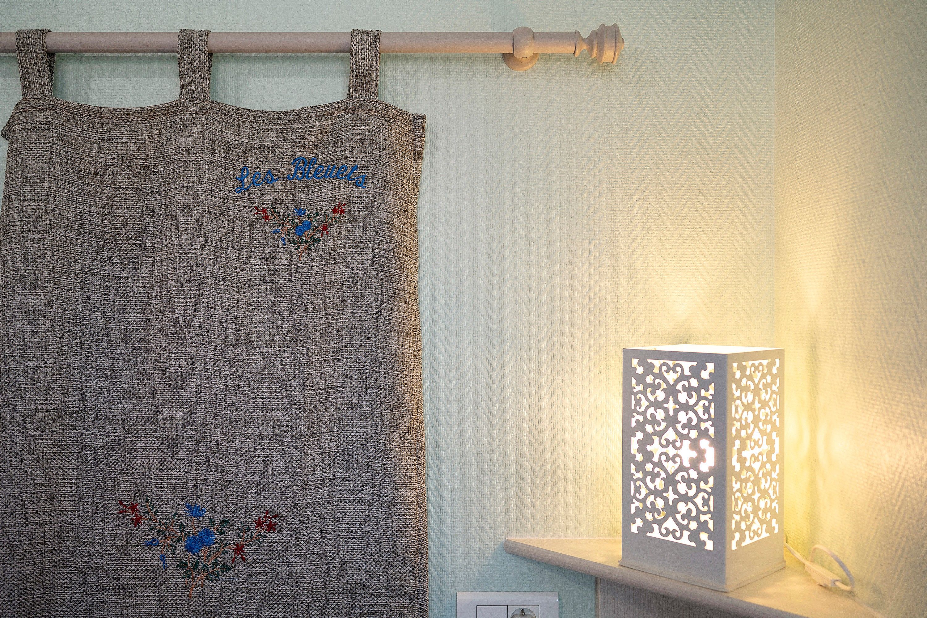A curtain with embroidered text and a decorative pattern, a wall-mounted light fixture, and a shelf with a white object.