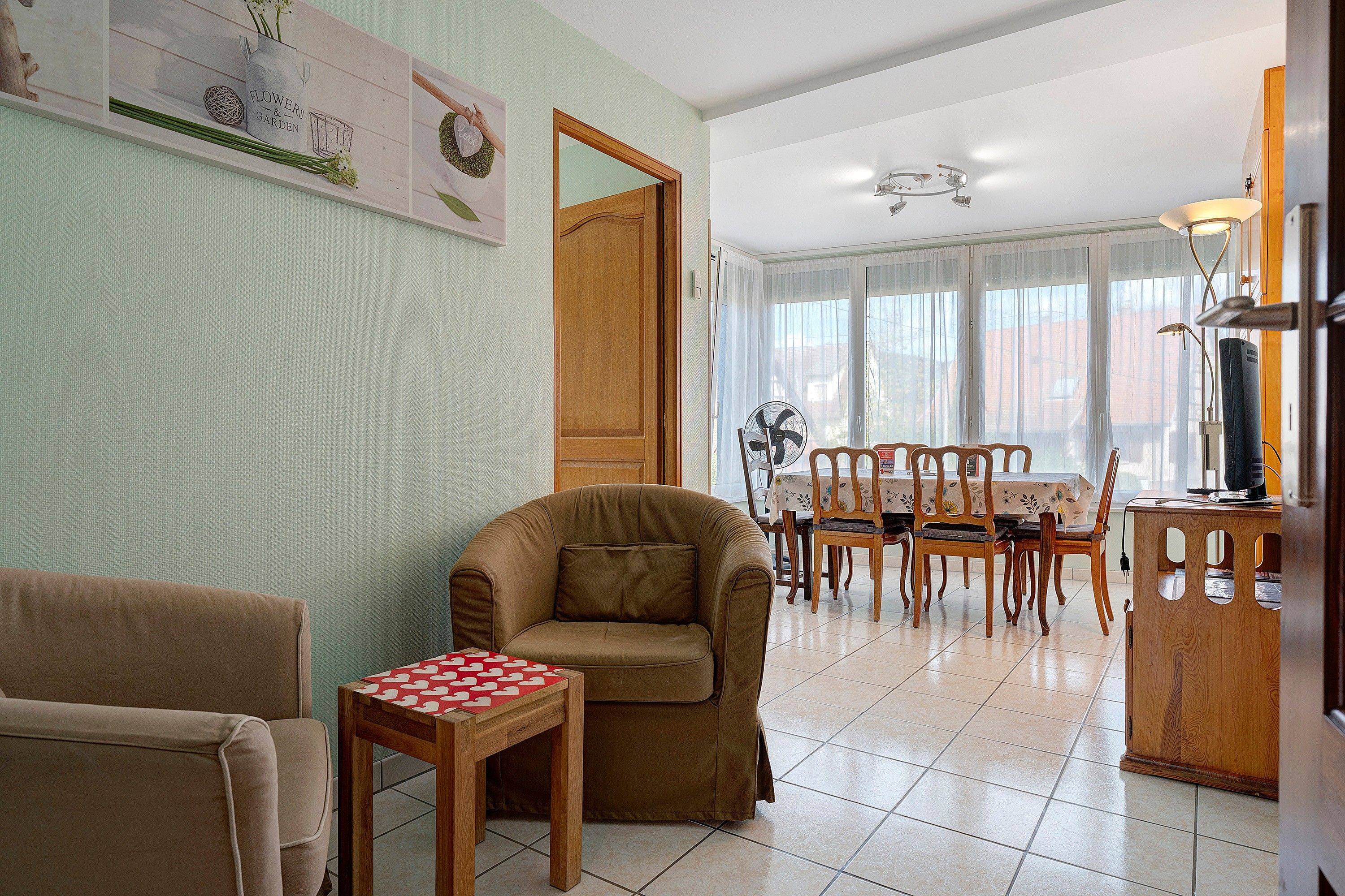A living room with a brown sofa, a wooden table with a red and white patterned tablecloth, and a dining area with wooden chairs and a table.