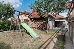 Een achtertuin met een houten schommel, een groene glijbaan, een stapel houtblokken en een terras met tuinmeubilair.