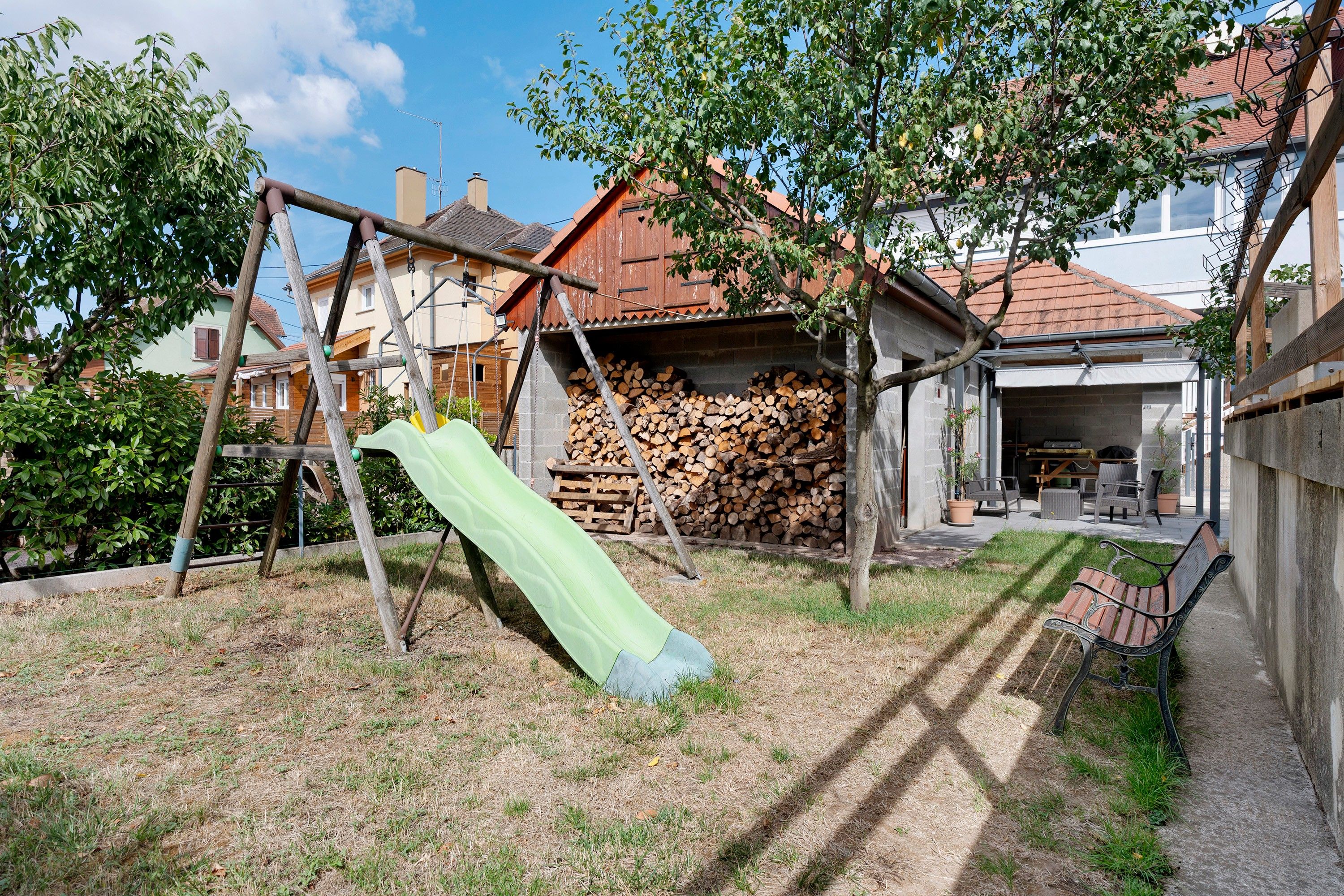 A backyard with a wooden swing set, a green slide, a stack of firewood, and a barbecue area.