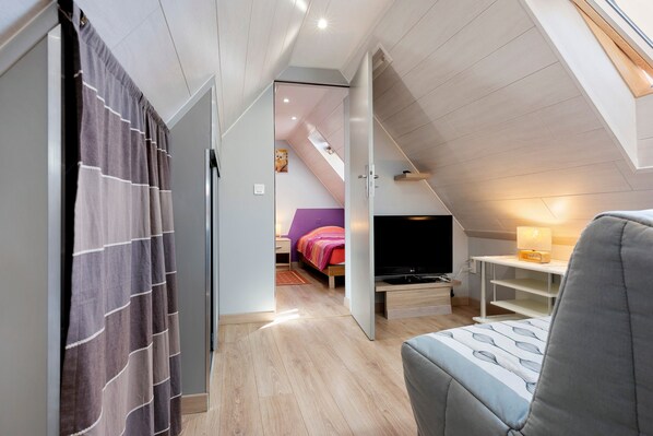 Une chambre à coucher moderne avec un plafond en pente, un lit recouvert d’une couverture colorée, une télévision et un bureau avec une lampe.