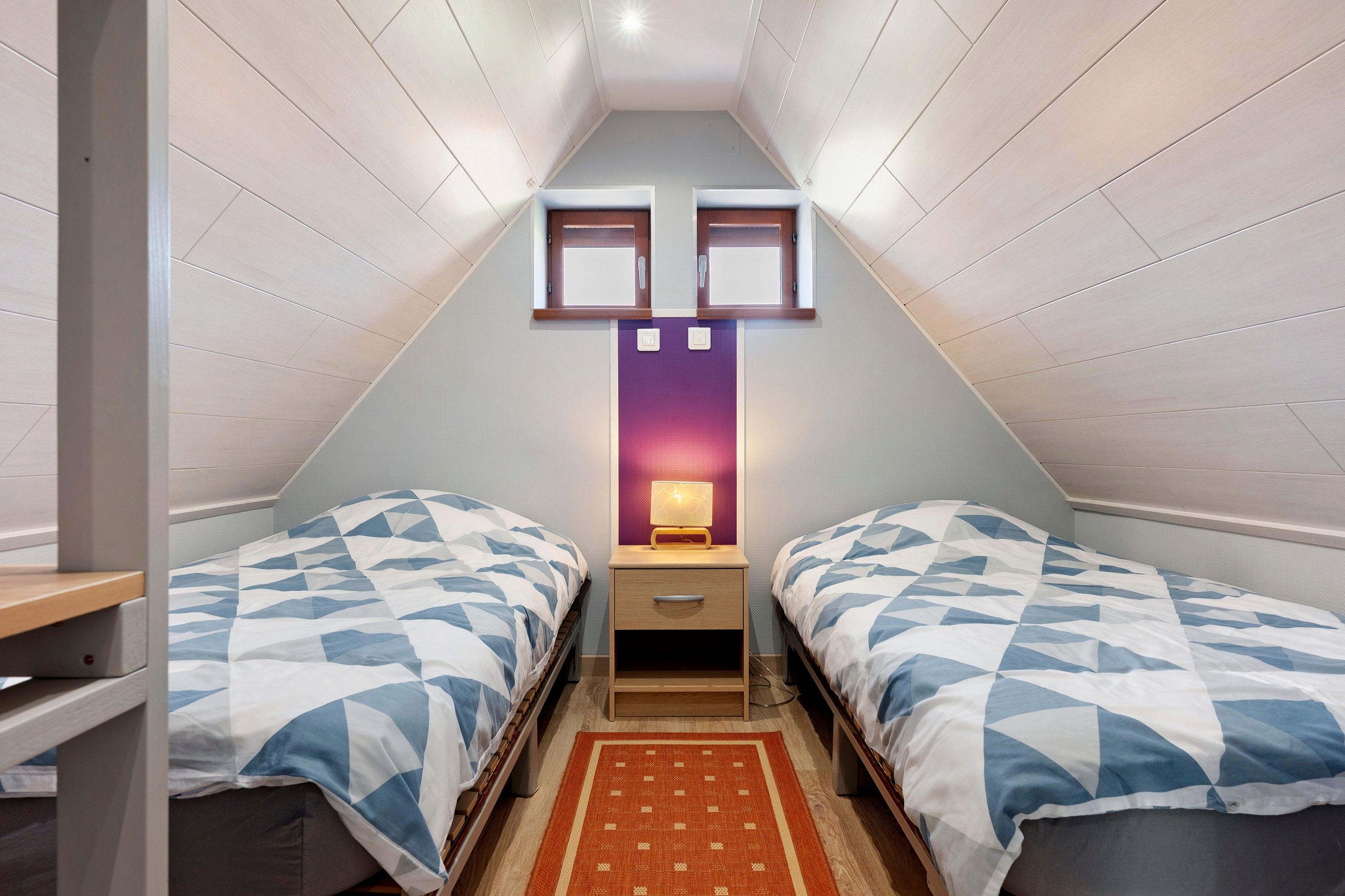 A small, triangular attic bedroom with two beds, a nightstand, a lamp, and a red rug.