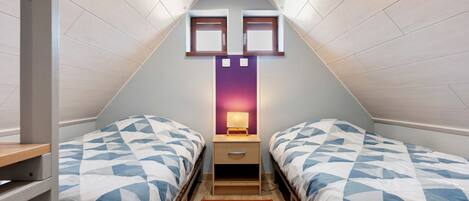 A small, triangular attic bedroom with two beds, a nightstand, a lamp, and a red rug.