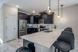 A modern kitchen with dark cabinets, stainless steel appliances, a large island with seating, and pendant lighting. - 8-bedroom house - ChampionsGate with pool. (Four Corners)