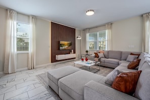 A modern living room with a sectional sofa, a flat-screen TV mounted on a wooden panel, a coffee table, and a patterned rug. - 8-bedroom house - ChampionsGate with pool. (Four Corners)