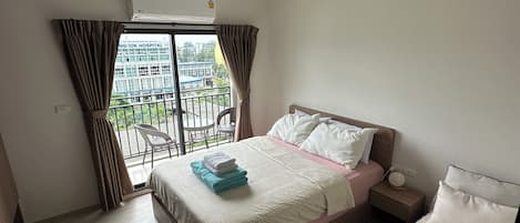 A hotel room with a bed, a bedside table, a nightstand, a window with curtains, and an air conditioning unit.