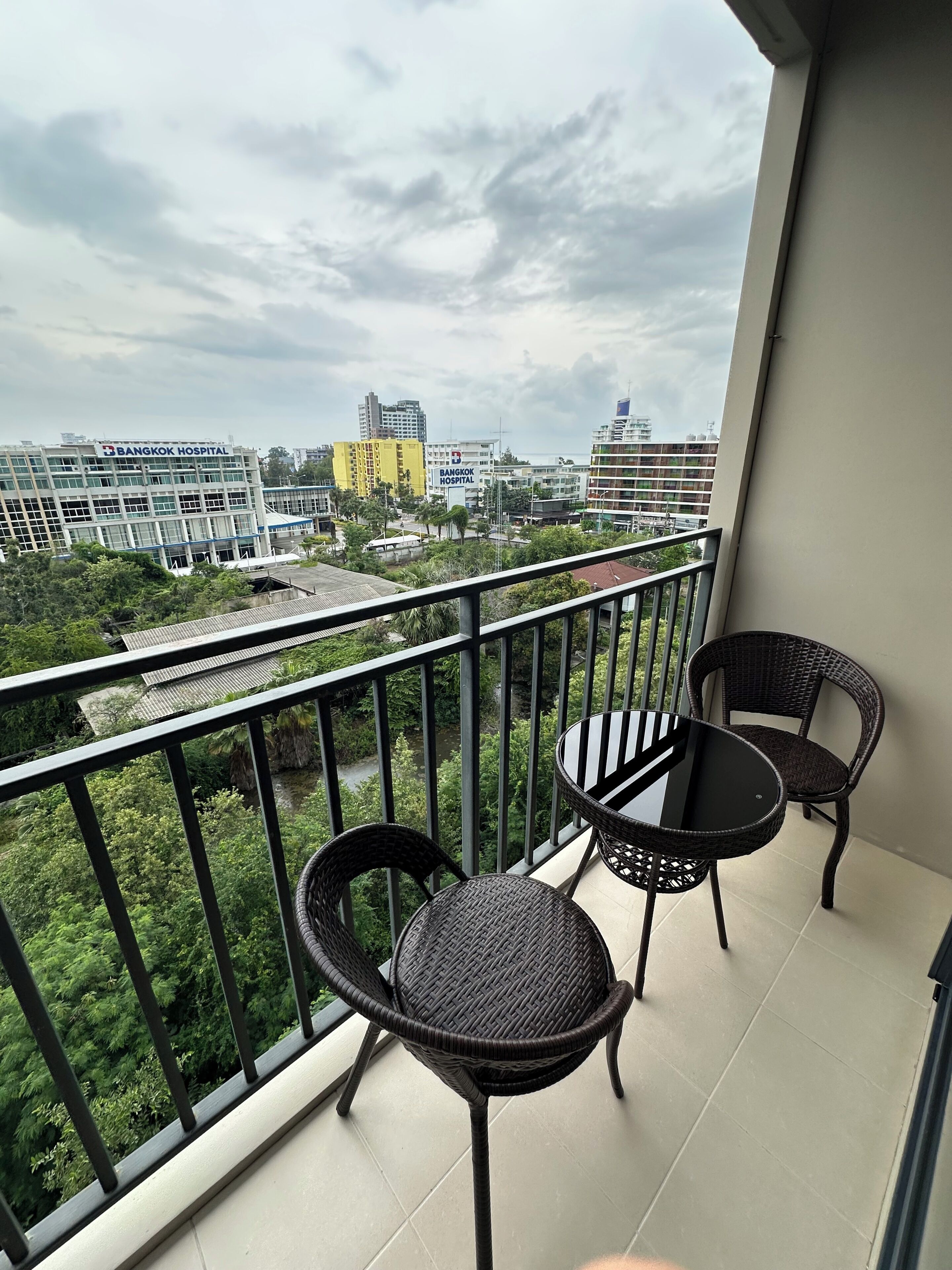A balcony with two wicker chairs and a table, overlooking a cityscape with buildings and greenery.