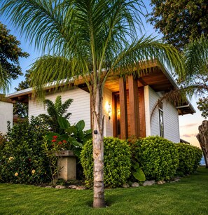 Una casa de fachada blanca, un porche de madera y un jardín cuidado con diversas plantas y una palmera.