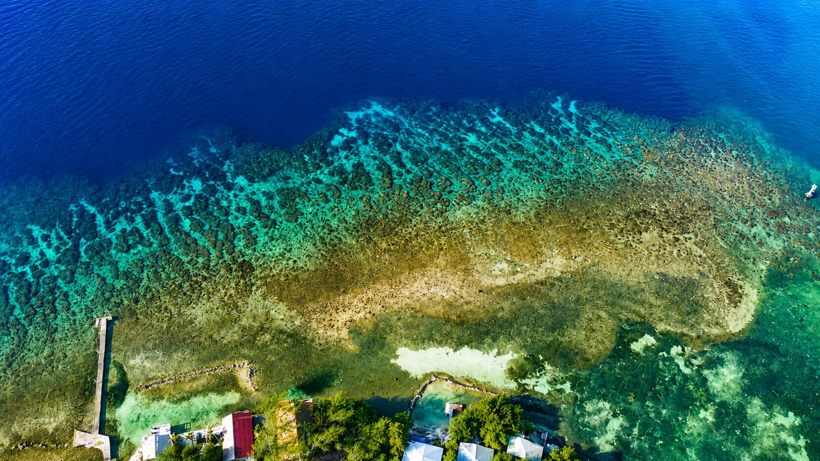 An aerial view of a coastal area with clear blue waters, a pier, and buildings at the shoreline.