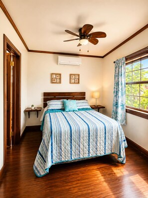 A bedroom with a bed, a ceiling fan, an air conditioning unit, a window with curtains, and a nightstand with a lamp. - Seas the Day-Oceanfront 2 Bedroom Water Access Only (Oakridge)
