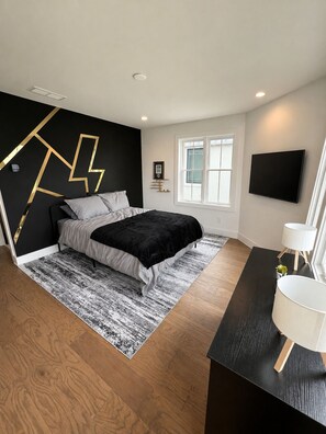 A modern bedroom with a bed, a black and white rug, a desk with a lamp, and a wall-mounted TV. - The Breakspear House - 4BR Trilith Stay for Production & Soccer in Fayetteville (Fayetteville)